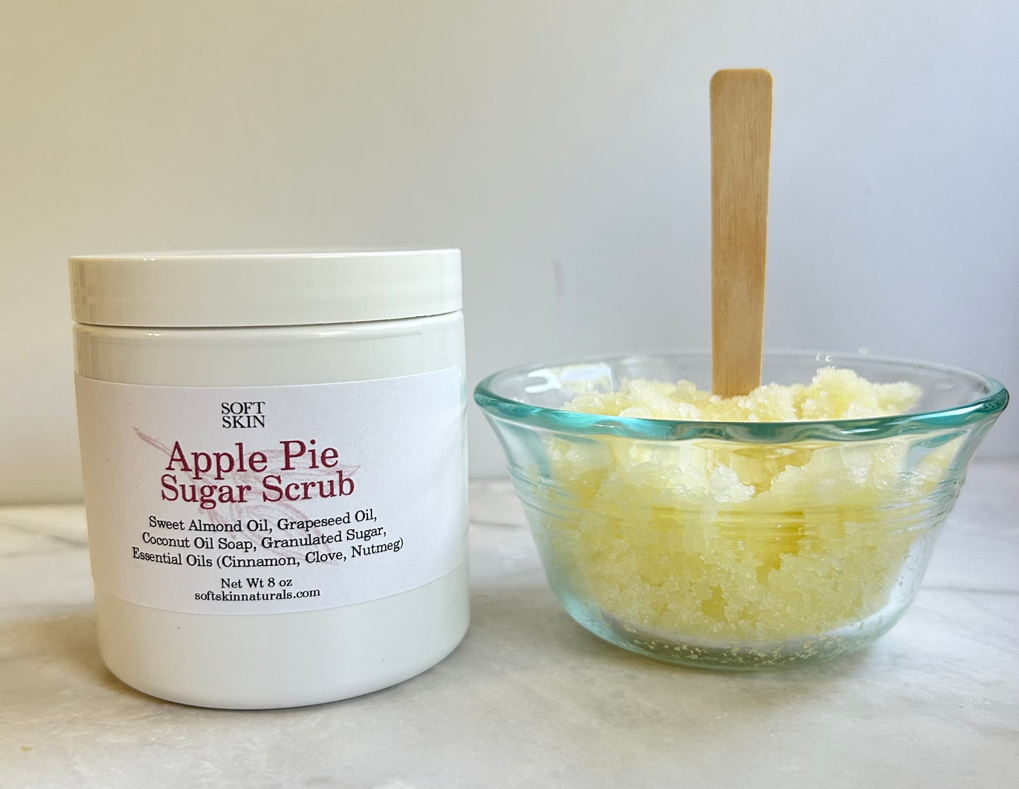 A white jar of Soft Skin Apple Pie Sugar Scrub next to a small clear glass bowl filled with pale yellow sugar scrub and a wooden spoon, set on a light marble surface against a white background.