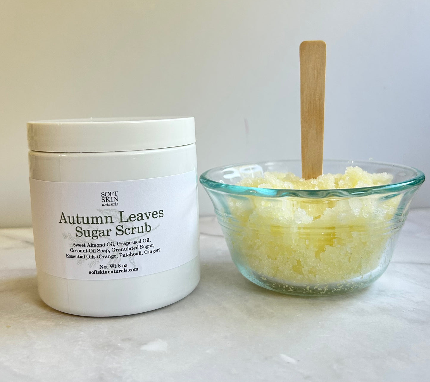 A white jar of Autumn Leaves sugar scrub stands next to a small clear glass bowl filled with the pale yellow scrub and a wooden applicator stick, set on a light marble surface against a neutral background.