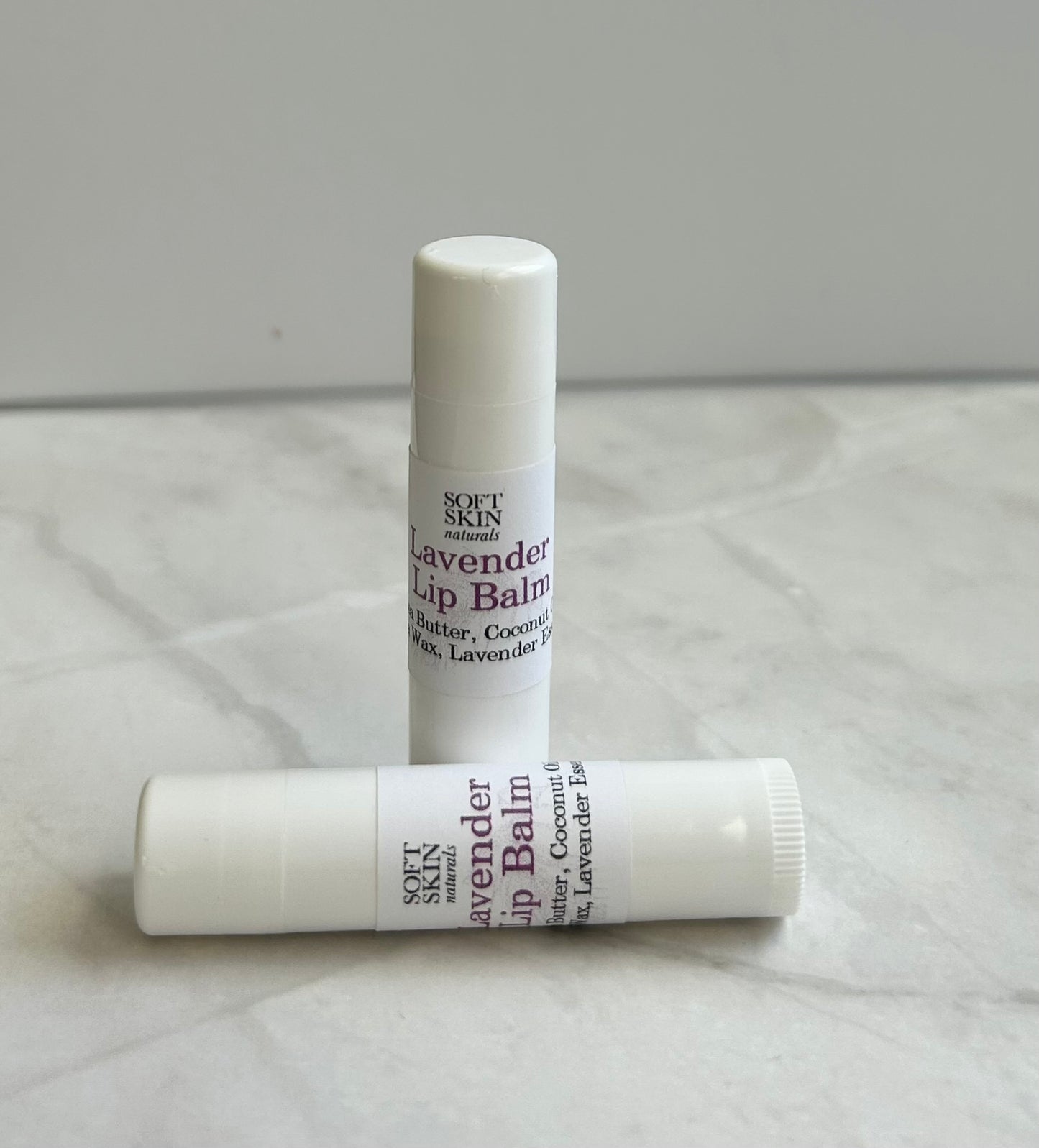 Two lavender lip balms on a textured surface with a neutral background