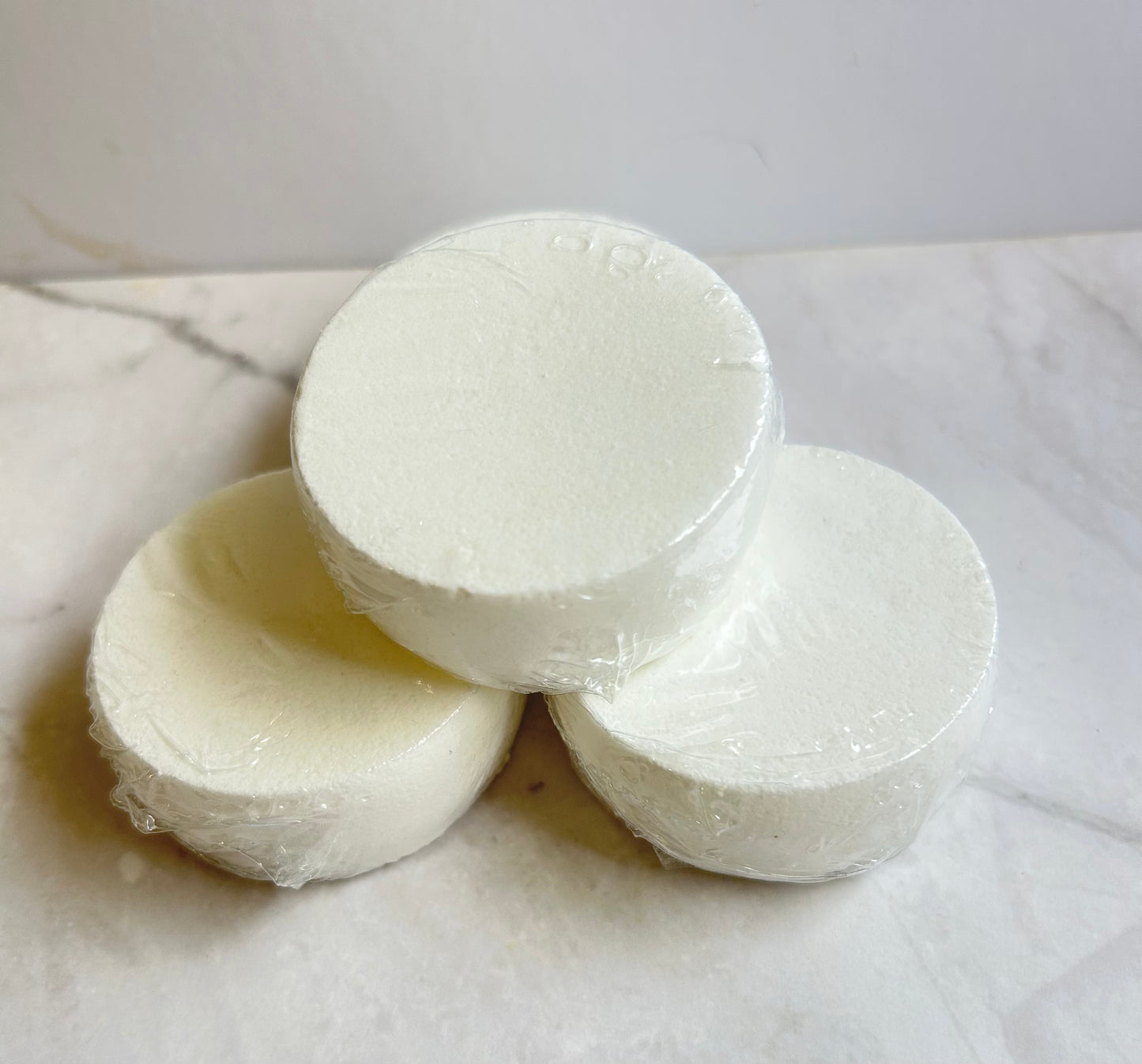 Three round white soap bars wrapped in clear plastic, stacked in a pyramid arrangement on a light marble surface.