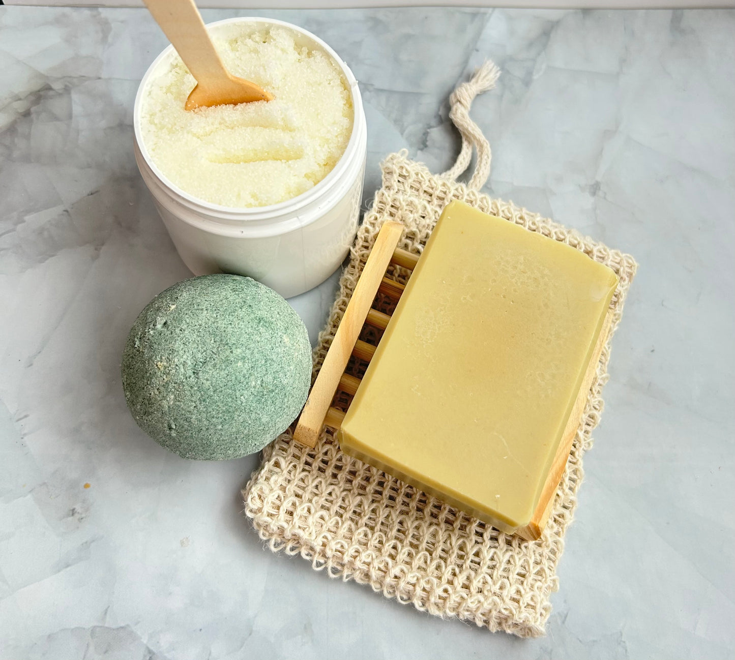 Bar of soap on a textured mat with a jar of bath salts and a green bath bomb on a marble surface.