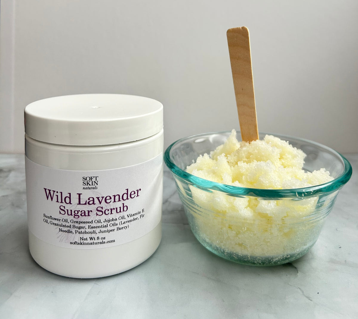 White container of Wild Lavender Sugar Scrub next to a glass bowl filled with sugar scrub on a marble surface.