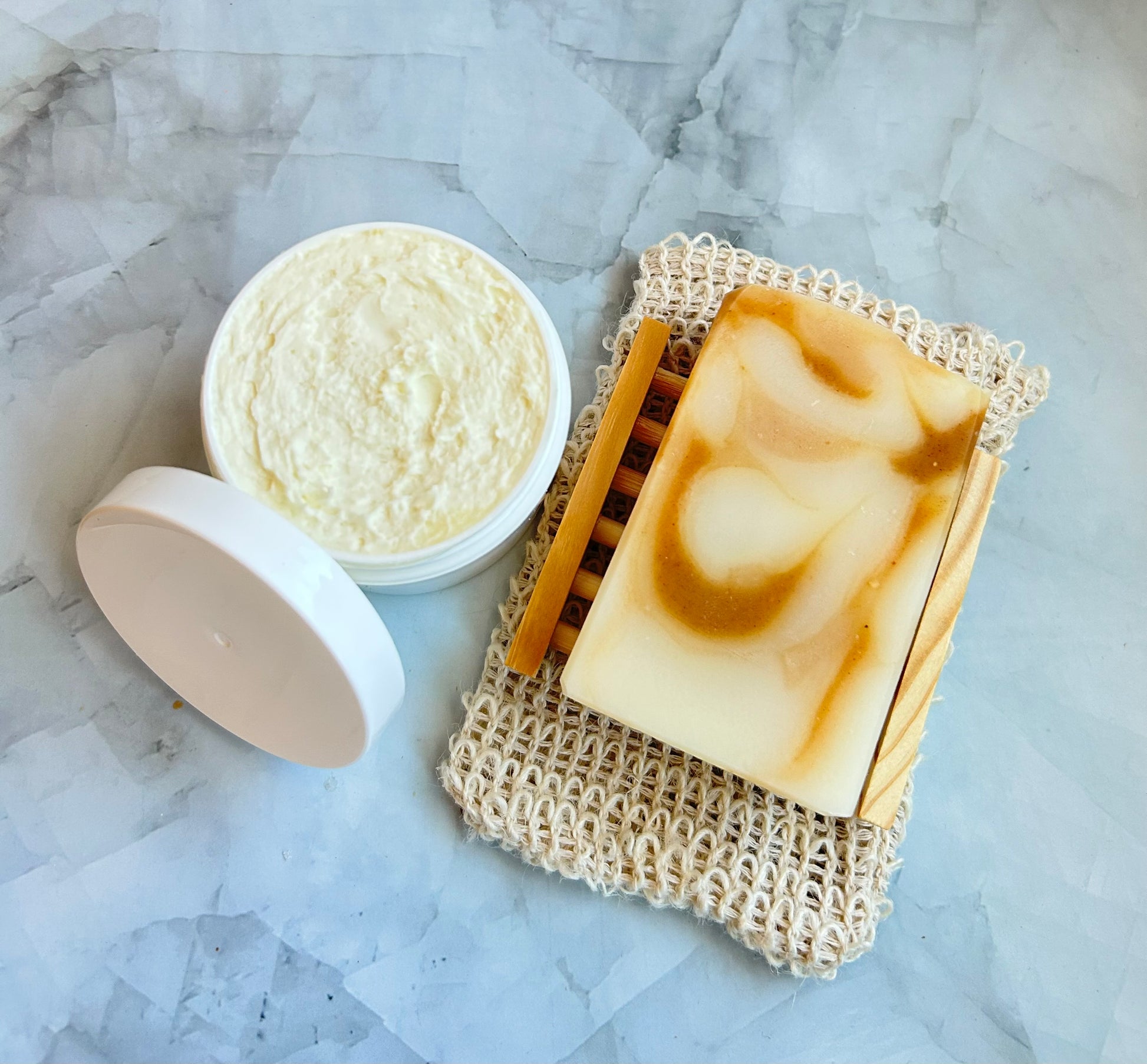 Bar of soap on a textured soap dish with a white container of body butter on a marble background