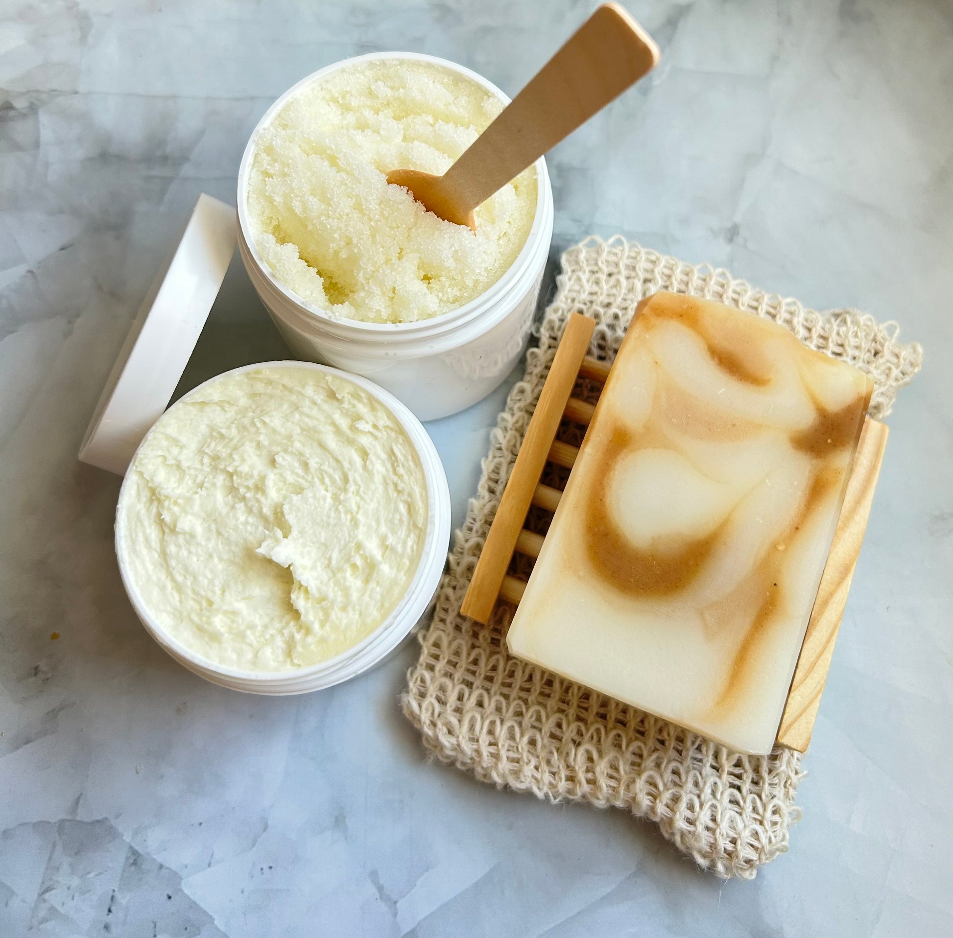 Container of scrub, jar of cream, and a bar of soap on a textured surface