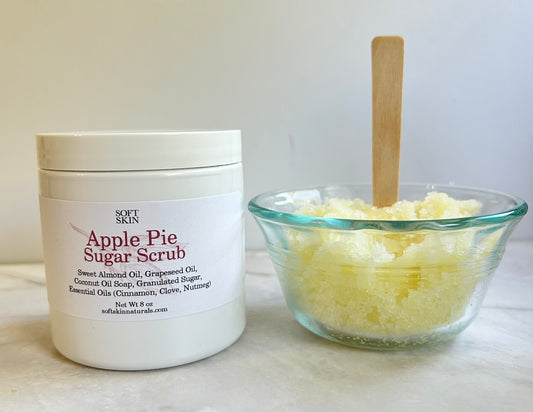 A white jar of Soft Skin Apple Pie Sugar Scrub next to a small clear glass bowl filled with pale yellow sugar scrub and a wooden spoon, set on a light marble surface against a white background.