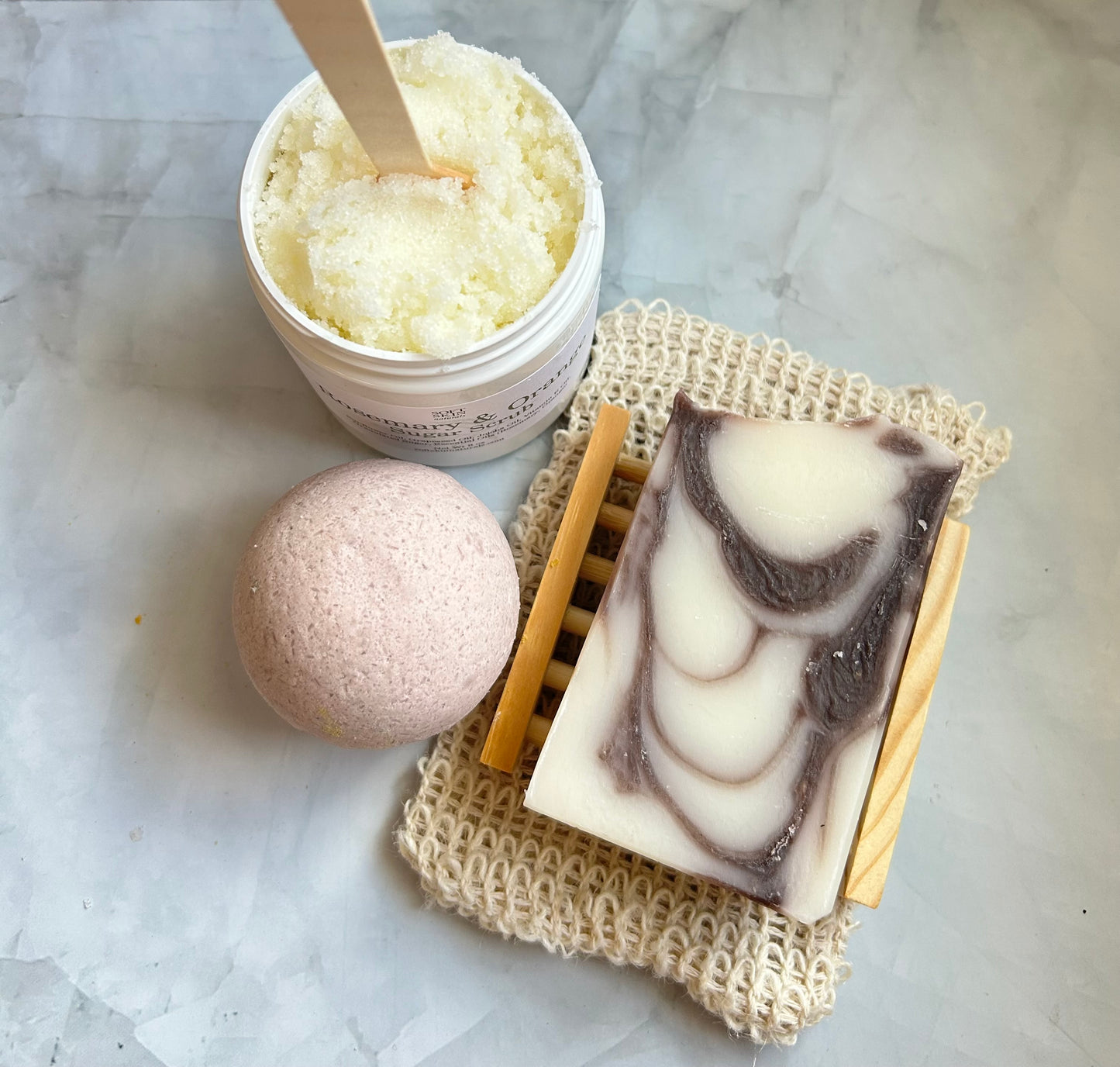 Bath bomb, container of bath scrub, and soap with a wooden spoon on a marble surface
