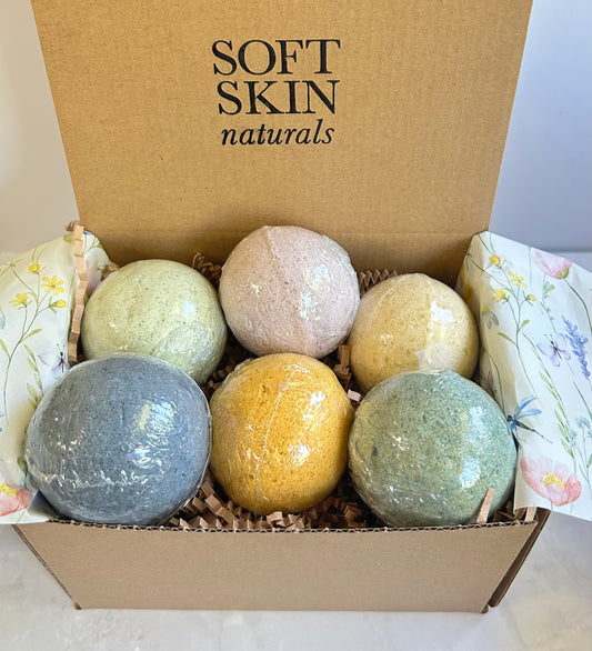 Six bath bombs in various earthy colors arranged in a cardboard gift box with floral tissue paper.
