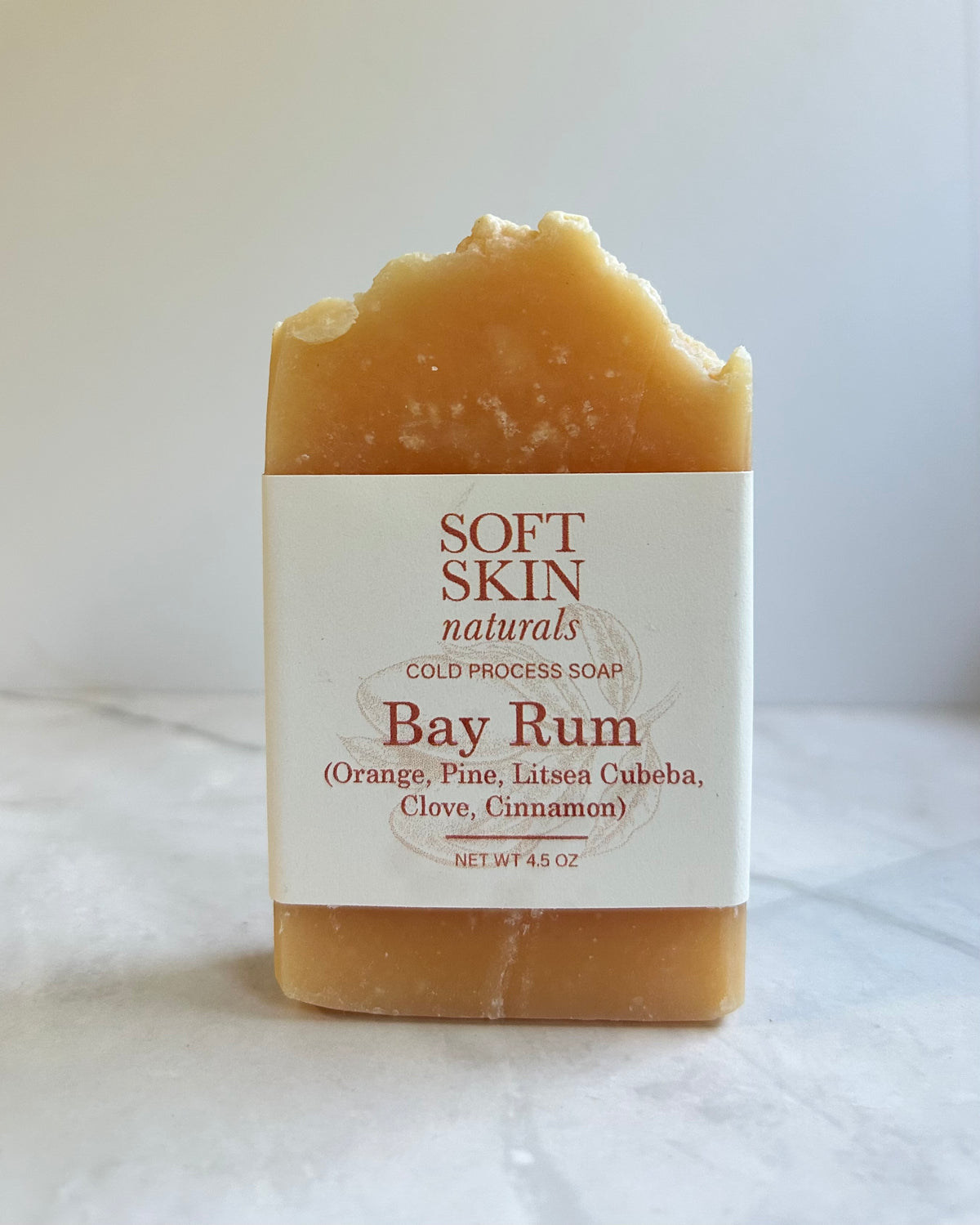 A rectangular bar of Soft Skin Naturals Bay Rum cold process soap, featuring a rustic, broken top edge. The soap is a warm golden-brown color with a white paper label wrapped around the center, set against a neutral light gray background.