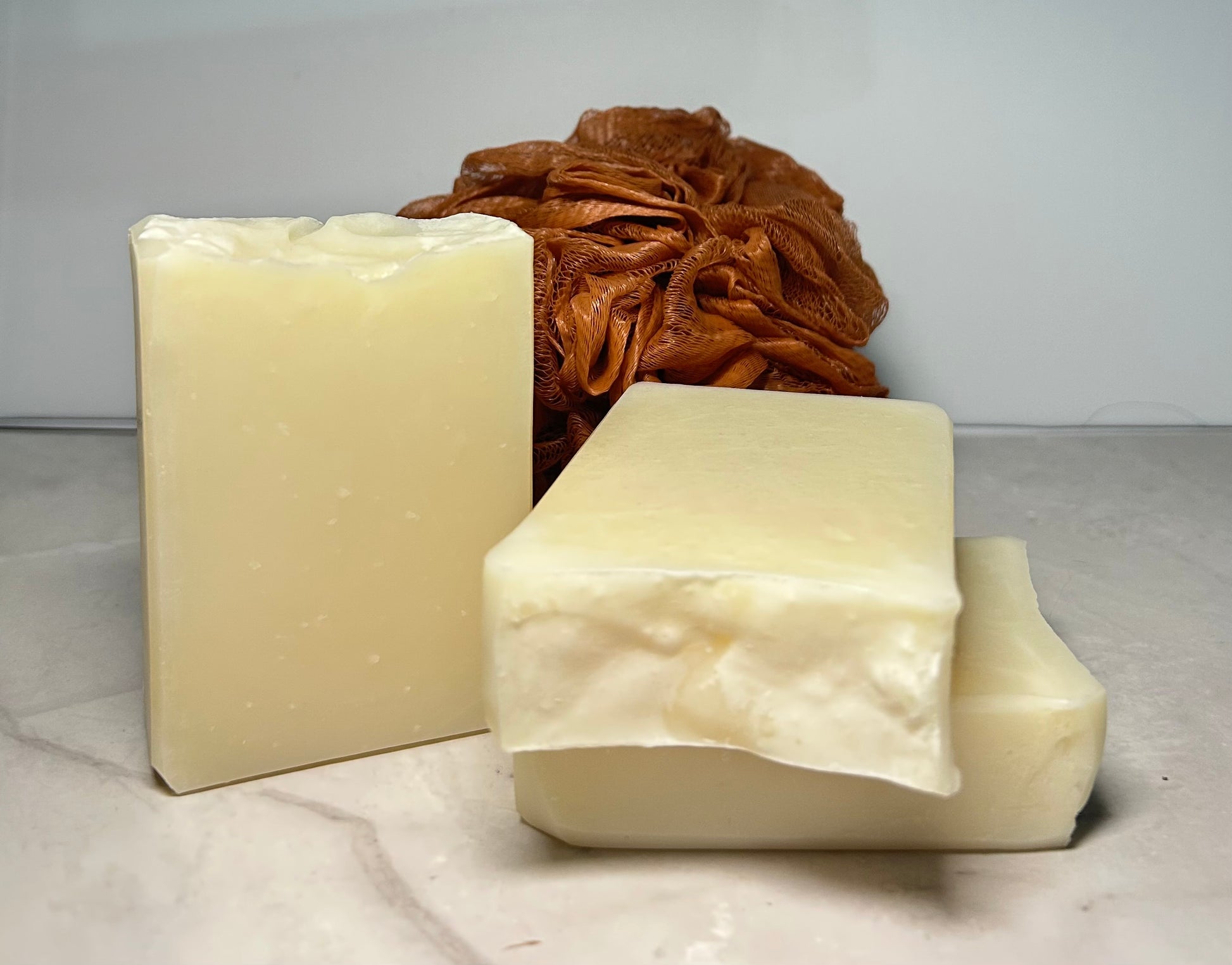 Two bars of soap and a brown loofah on a light gray surface with a white background