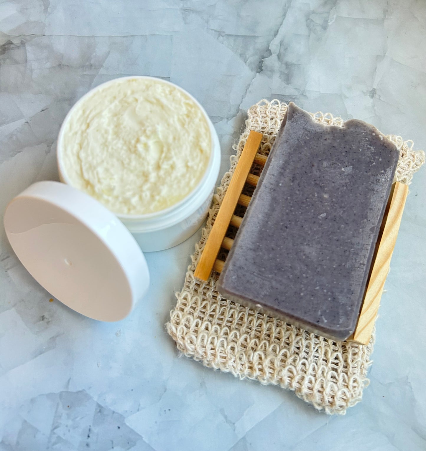 Bar of soap on a soap dish with a container of body butter on a marble surface