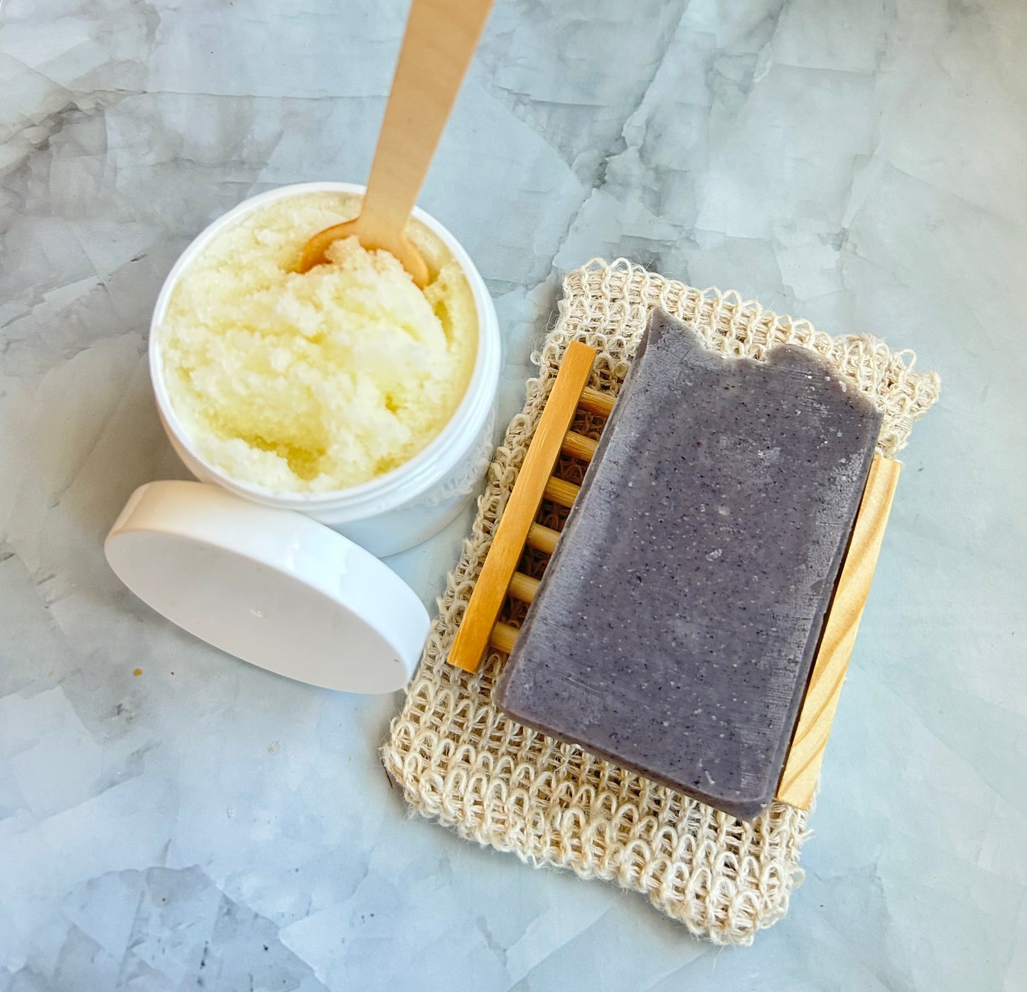 Bar of soap on a natural fiber soap dish with a container of sugar scrub on a marble surface.