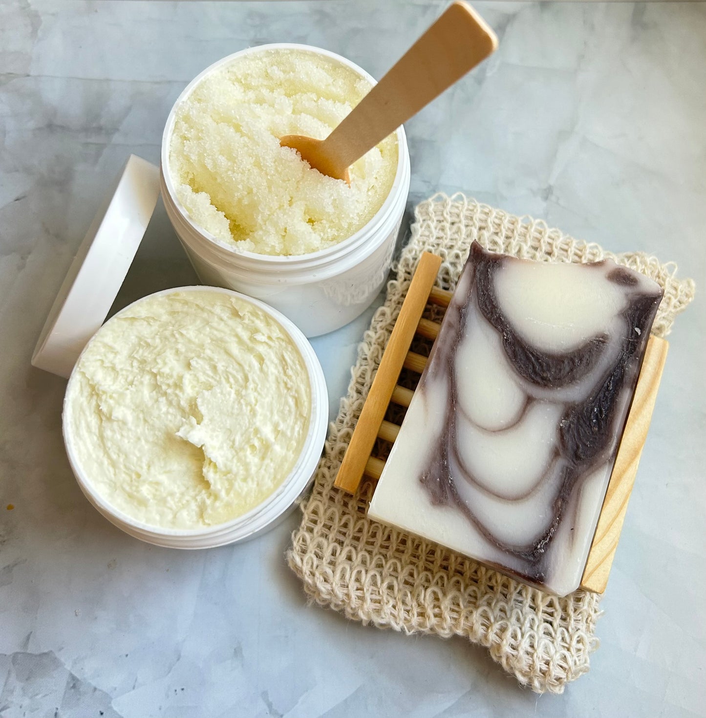 Containers of scrub, jar of cream, a wooden spatula, and a bar of soap on a textured surface.