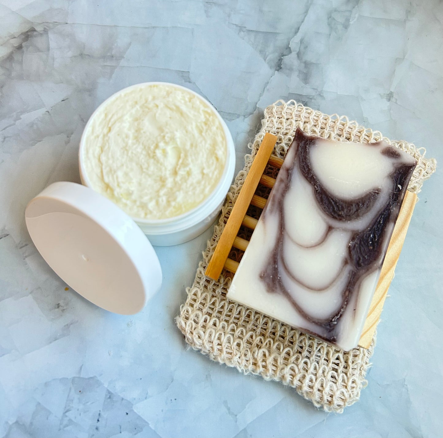 Bar of soap with a marbled pattern on a natural sponge, next to a container of body butter on a marble surface.