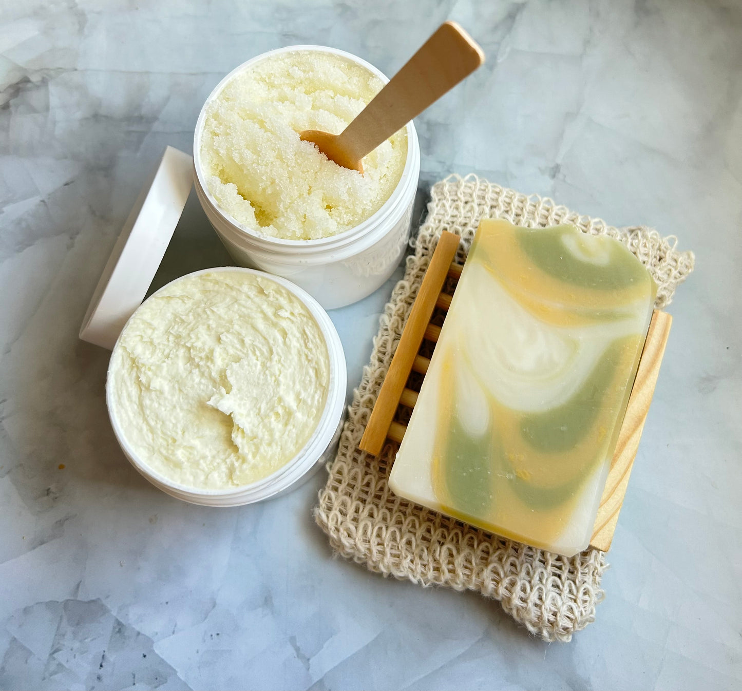 Container of scrub, jar of cream,  and a bar of soap on a marble surface