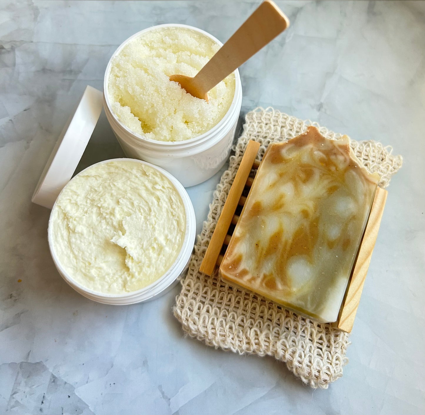 Container of scrub, jar of cream, a bar of soap on a wooden soap dish, and a wooden spoon on a marble surface.