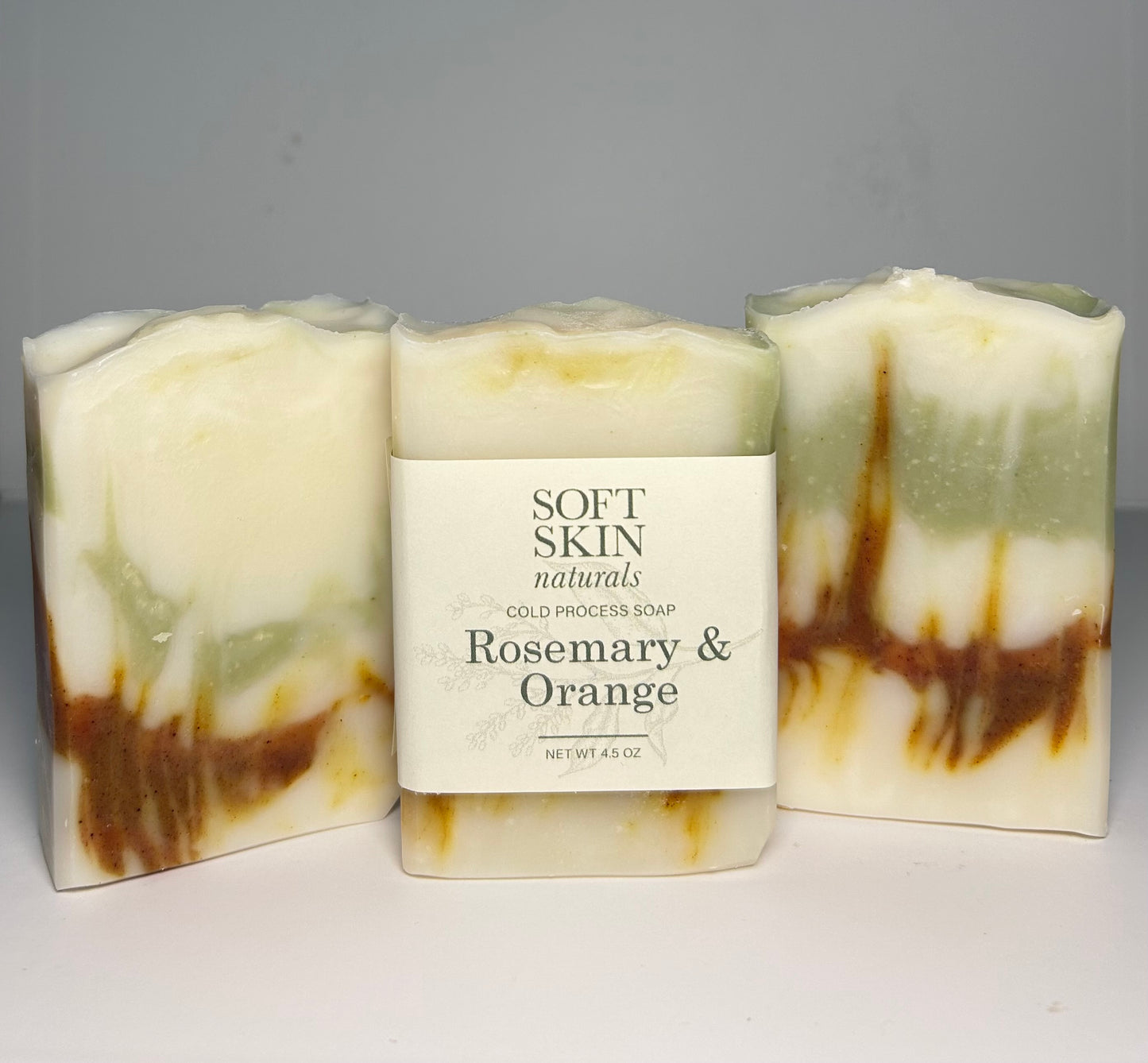 Three bars of soap with a label reading Soft Skin Naturals Rosemary & Orange on a gray background.