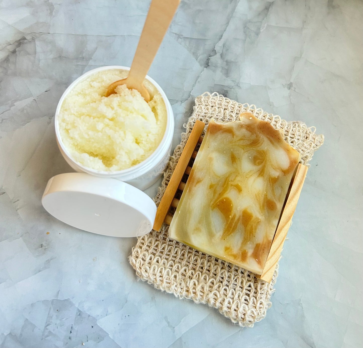 Bar of soap on a textured soap dish next to a container of scrub on a marble surface