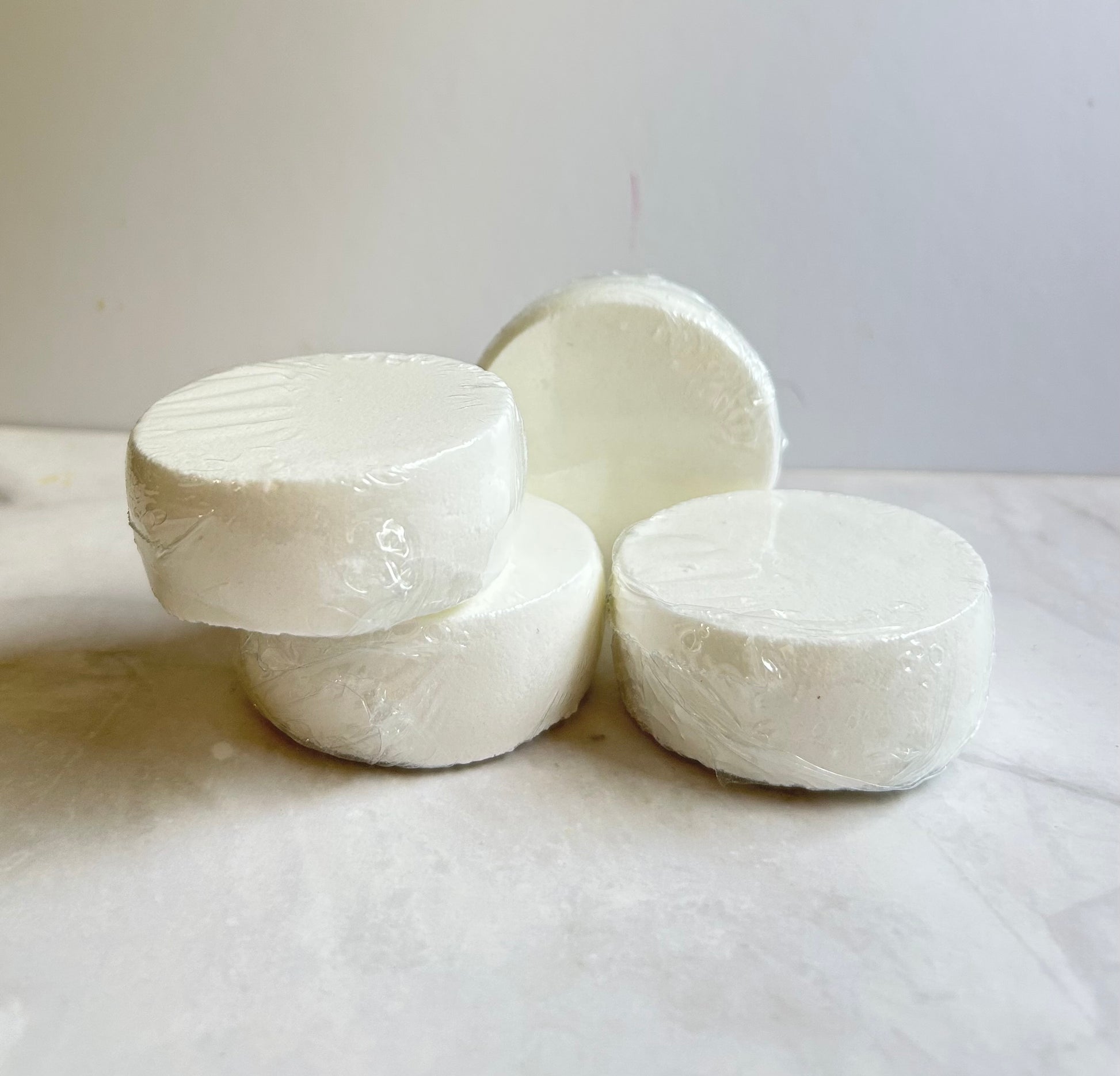Four round white soap bars wrapped in clear plastic, arranged on a light marble-like surface against a plain light gray background.