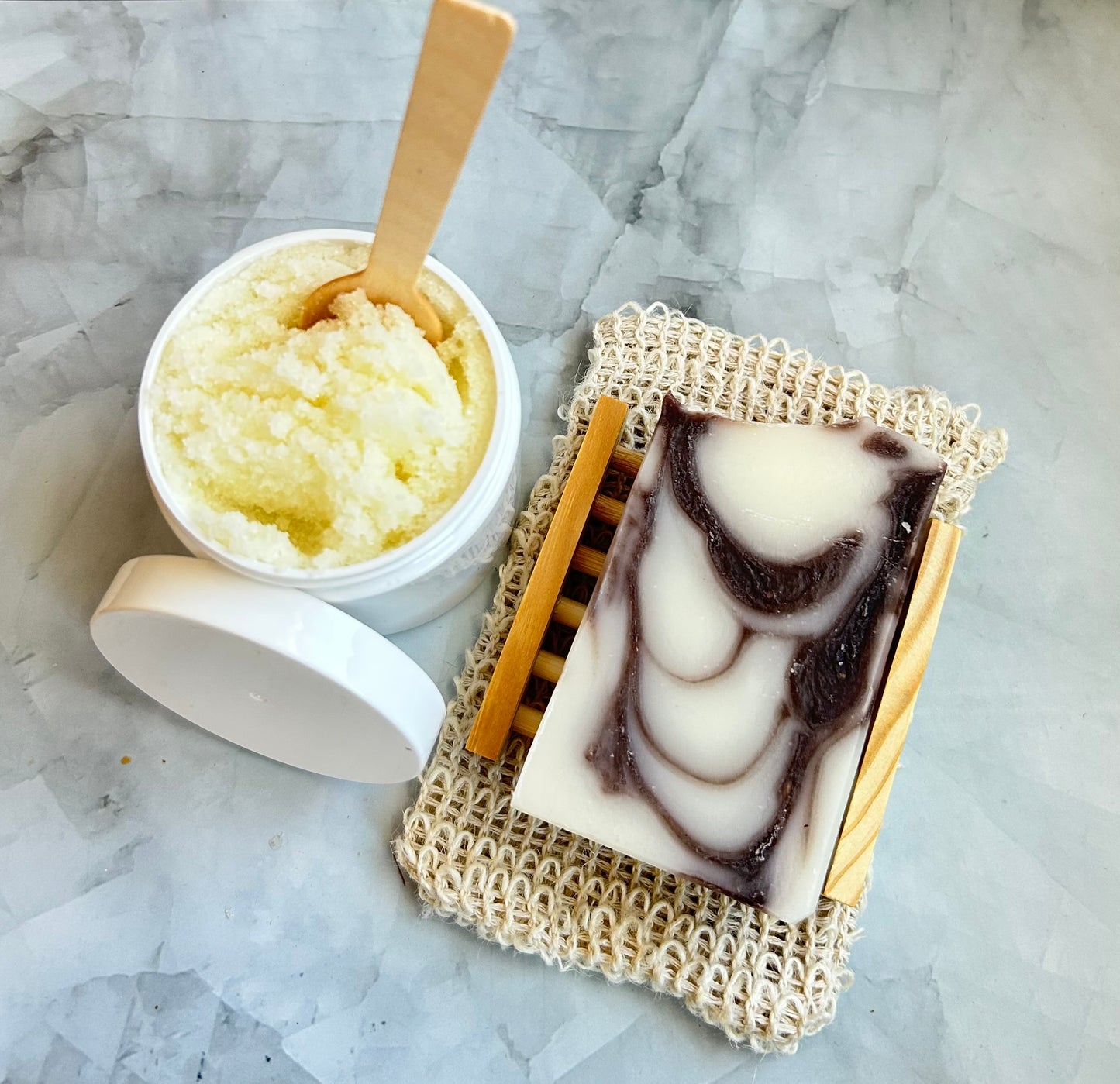 Marble-patterned soap dish with a bar of soap, a container of scrub, and a wooden spoon on a marble surface.