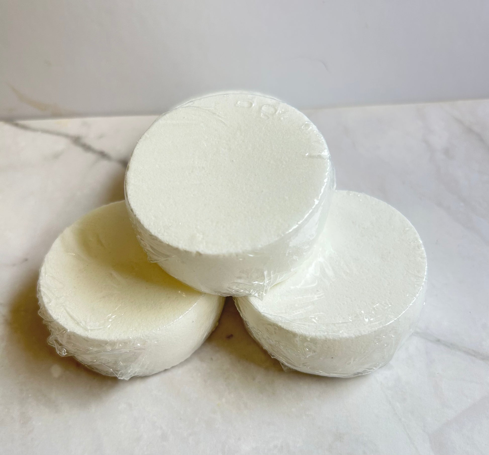 Three round white soap bars wrapped in clear plastic, stacked in a pyramid arrangement on a light marble surface.