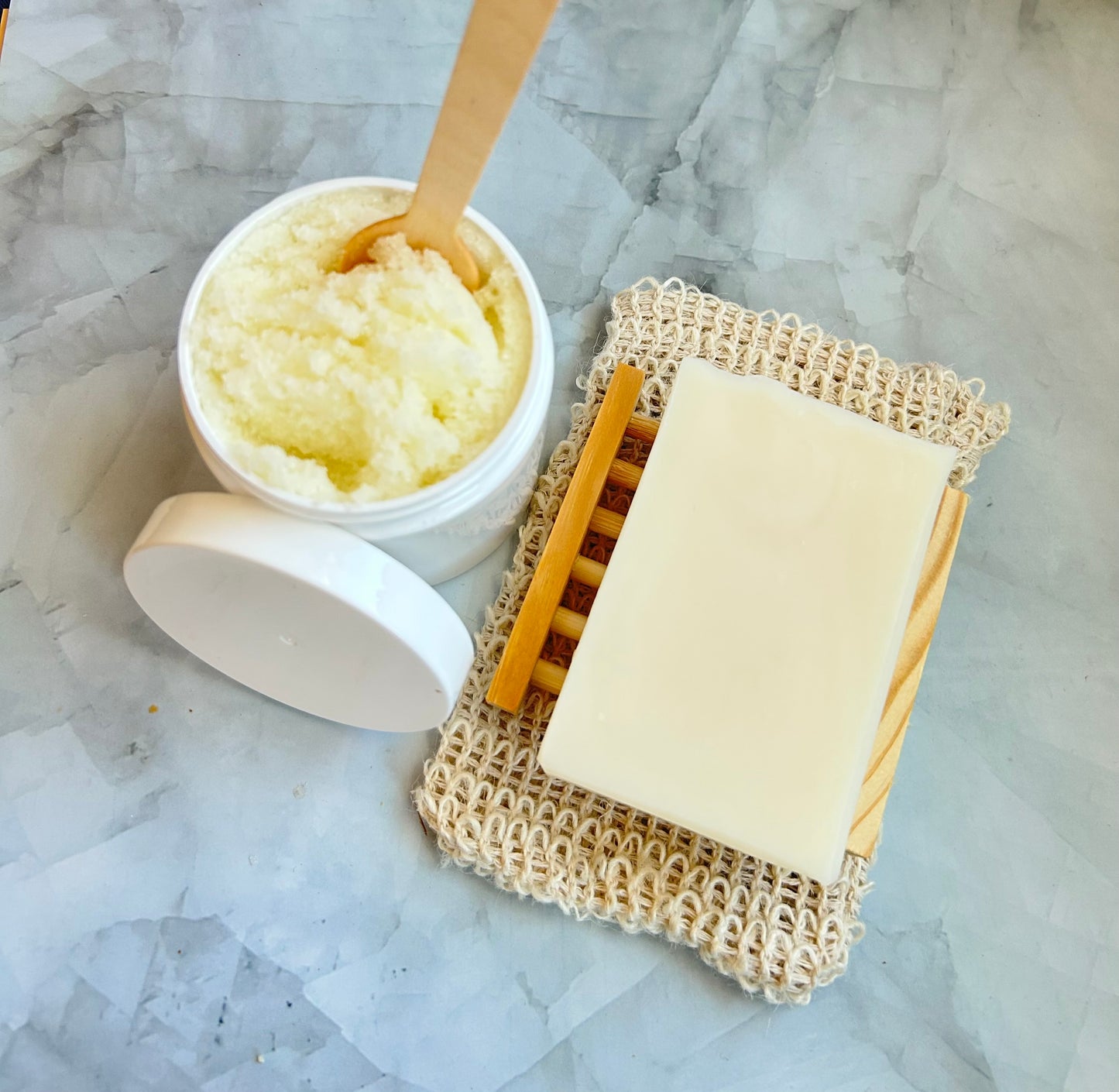 Bar of soap on a natural soap dish with a jar of sugar scrub on a marble surface