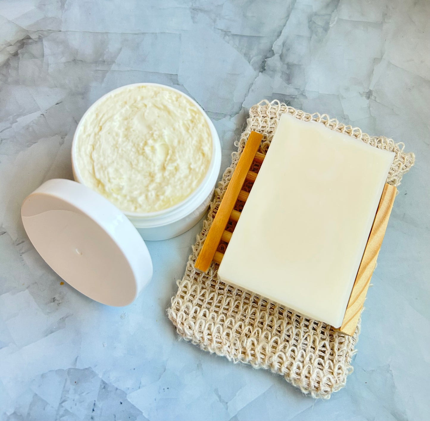 Bar of soap on a natural fiber soap dish with a jar of body butter on a marble background