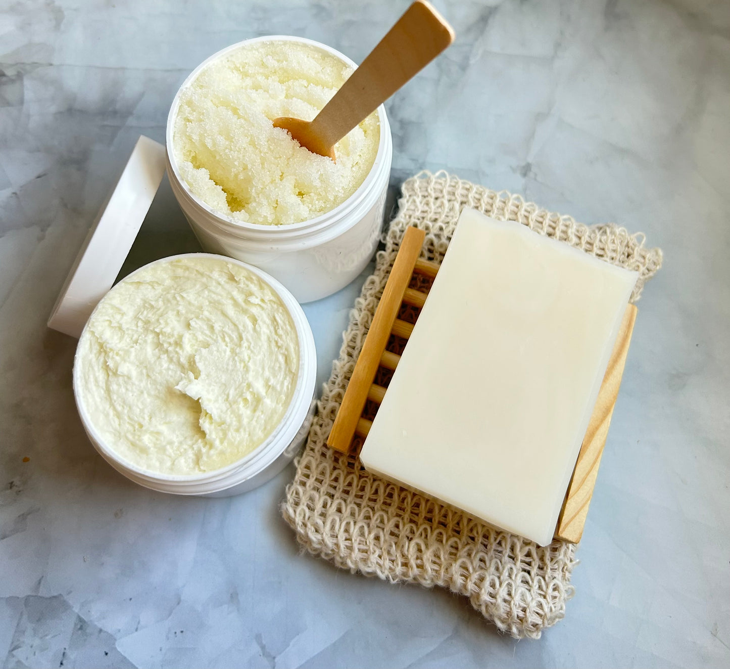 Container of scrub, jar of cream, a bar of soap on a soap dish, and a wooden spoon on a marble surface.