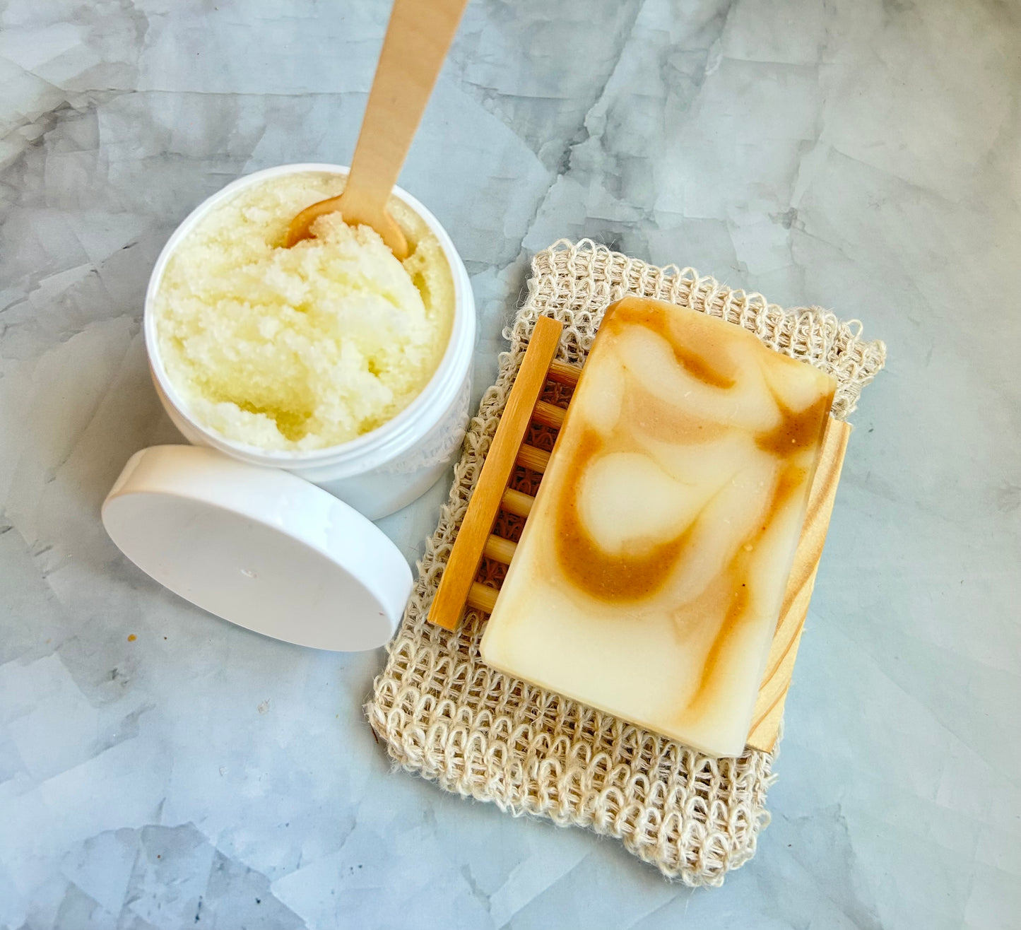 Bar of soap on a soap dish with a container of sugar scrub on a marble surface