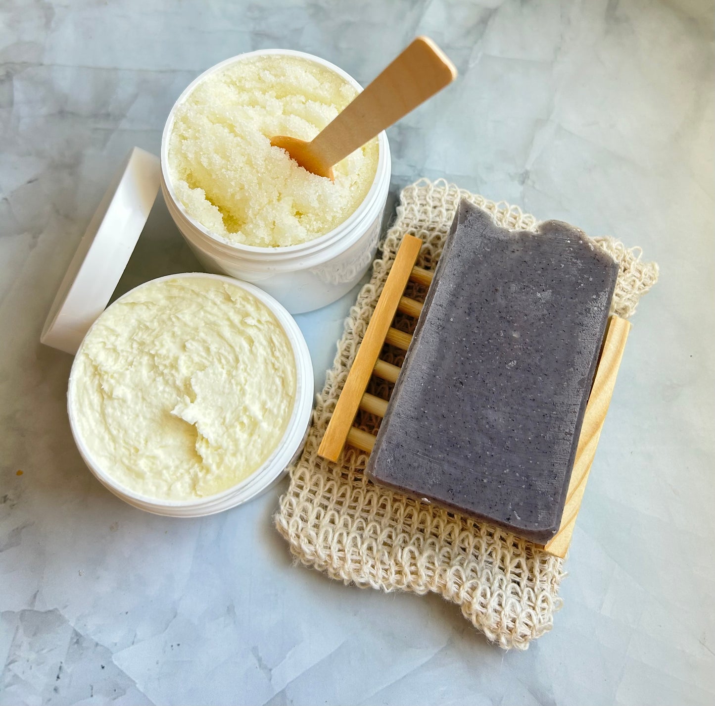 Container of scrub, jar of cream, a wooden spatula, a black bar of soap, and a beige soap dish on a marble surface.