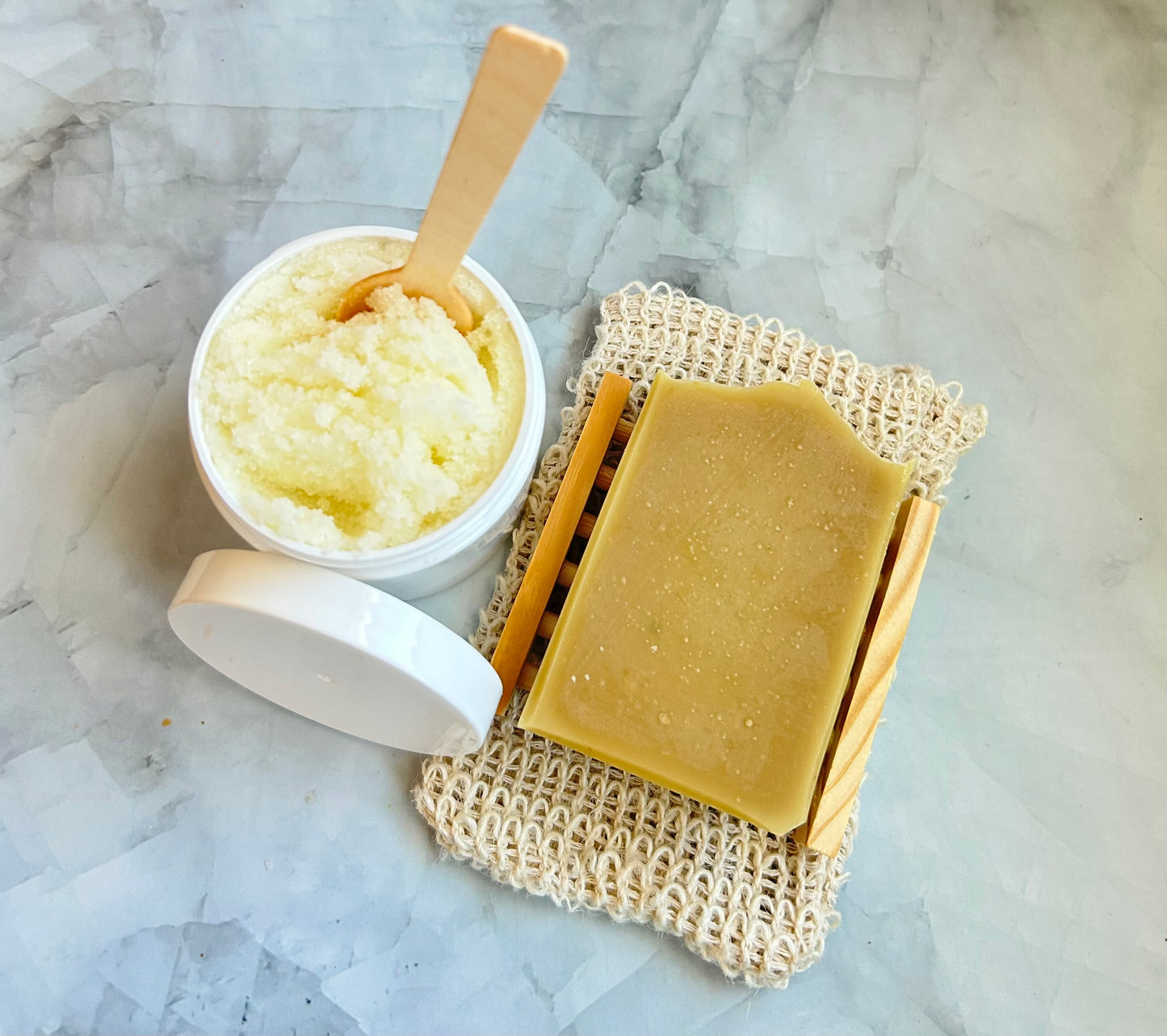 Bar of soap on a natural fiber soap dish next to a container of sugar scrub on a marble surface.