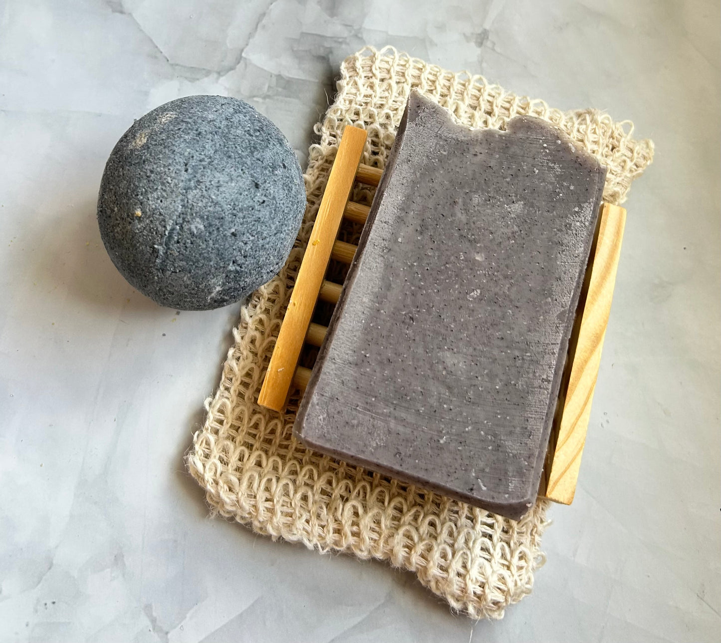 Bar of soap on a natural fiber soap dish with a wooden soap dish and a bath bomb on a marble surface.